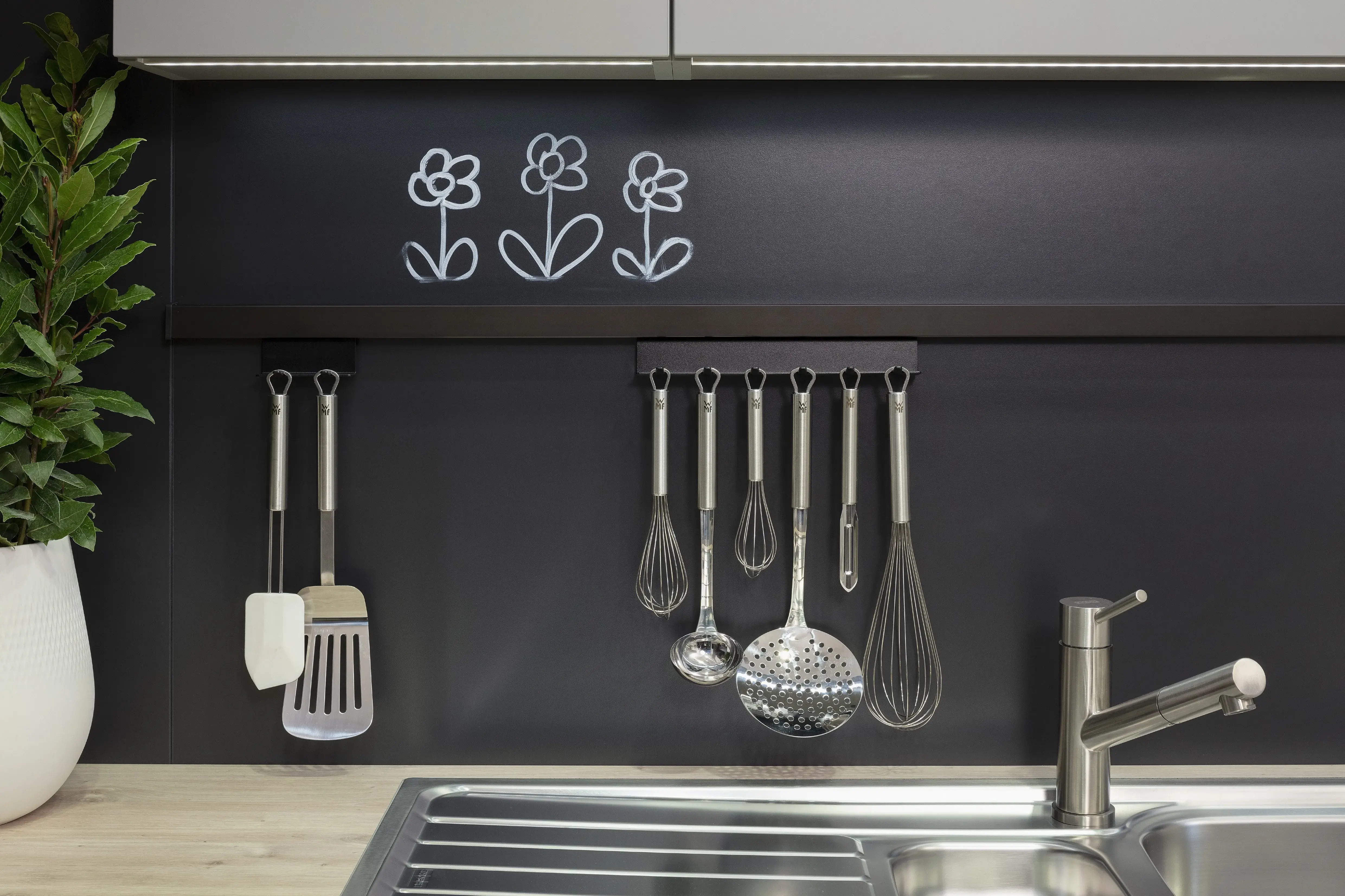 Küchenutensilien hängen an schwarzer Wand neben Spüle, oben weiße Blumenzeichnungen.